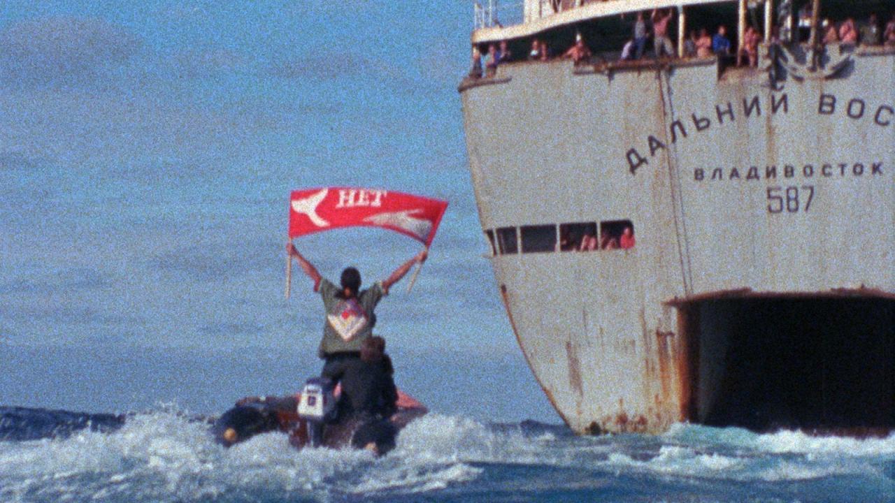 How To Change The World, Szenenbild: eine Figur steht auf ein Schlauchboot auf dem Meer und hält eine rote Fahne. Neben ist ein großer grauen Schiff.