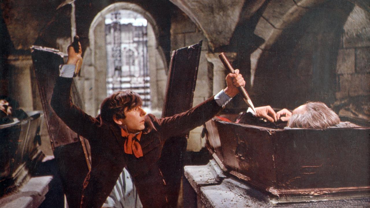 Szenenbild aus dem Film TANZ DER VAMPIRE (1967): In einer Gruft steht ein junger Mann neben einem Sarg. Während er mit der einen Hand einen Holzpflock auf die darin liegende Person richtet, holt er mit der anderen mit einem Hammer zum Schlag aus.