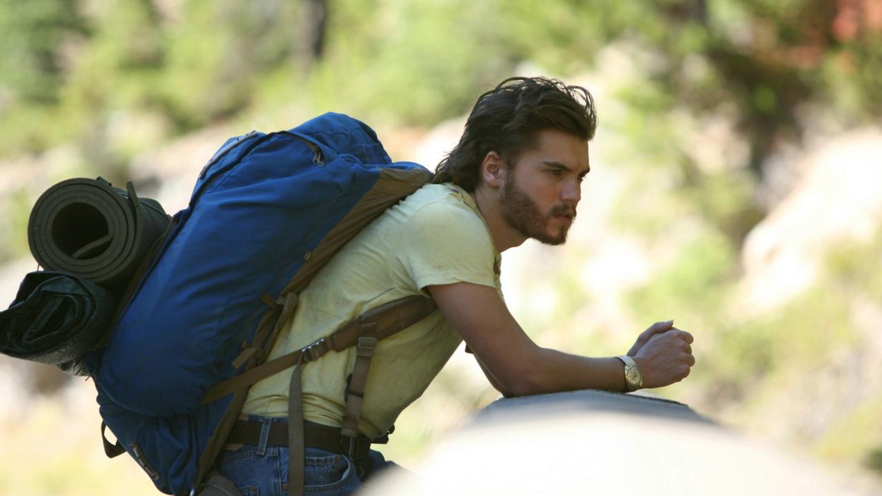 Into the wild, Szenenbild: ein junger Mann mit Bart, gelber T-shirt und Wanderrucksack lehnt sich an einem Gelände. Im Hintergrund ist Natur.