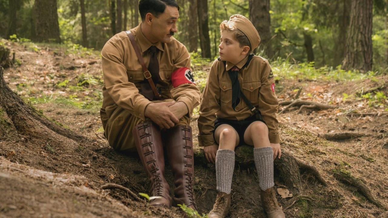 Jojo Rabbit, Szenenbild: Ein Mann, der aussieht wie Adolf Hitler, sitzt im Wald neben einem kleinen Jungen und hört ihm zu.