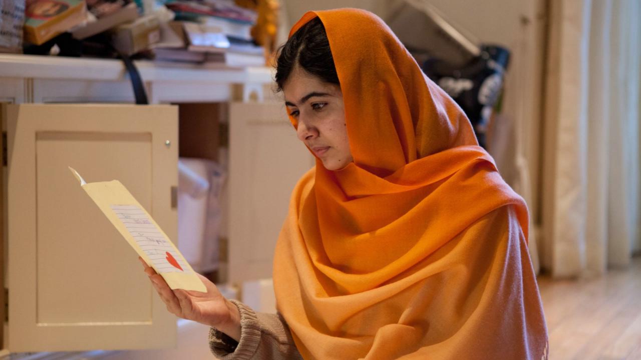 Malala – Ihr Recht auf Bildung, Szenenbild: ein junges Mädchen mit orangenen Kopftuch sitzt auf dem Boden und liest von einer Karte, die sie in der Hand hält.