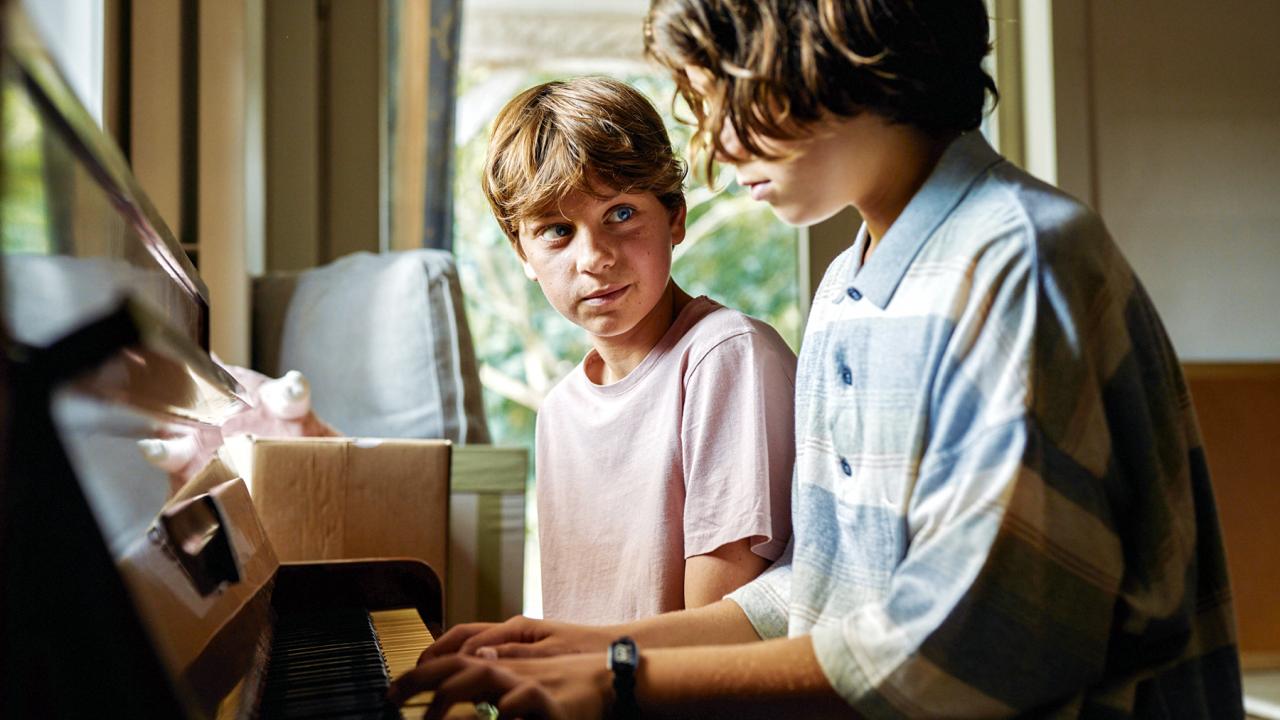 Filmstill aus dem Coming-of-Age-Film YOUNG HEARTS: Ein Junge im Teeanger-Alter sitzt am einem Klavier und spielt. Neben ihm sitzt ein anderer Junge und schaut ihn an.