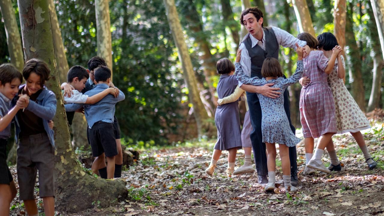 ilmbild aus dem Drama DER LEHRER, DER UNS DAS MEER VERSPRACH: Ein Mann und mehrere Kinder, alle im Stil der 1940er gekleidet, tanzen paarweise zwischen Bäumen.