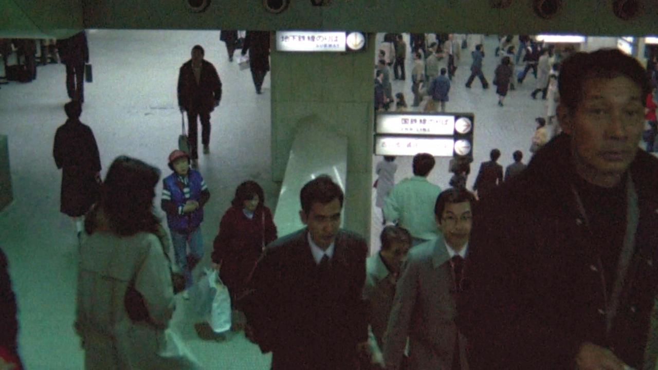 Filmstill aus SANS SOLEIL – UNSICHTBARE SONNE: Menschen gehen in einer Tokioer U-Bahnstation eine Treppe hoch. Das Bild ist dunkel, ein Mann schaut direkt in die Kamera.