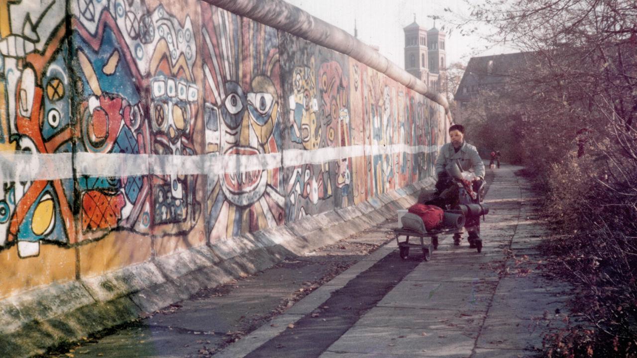 Striche ziehen, Szenenbild: eine Person mit einem Wagen voller Sachen läuft neben der Berliner Mauer, voller Graffiti. 