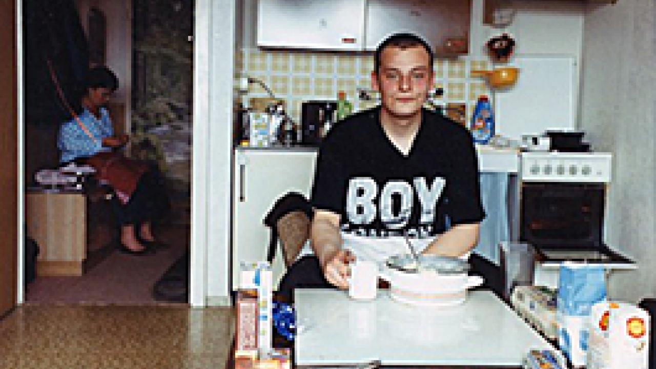 Stau – Jetzt geht's los, Szenebild: Ein junger Mann sitzt an einem Tisch in der Küche. Auf seinem T-Shirt steht "Boy". Im Hintergrund sitzt die Mutter in einem anderen Zimmer.