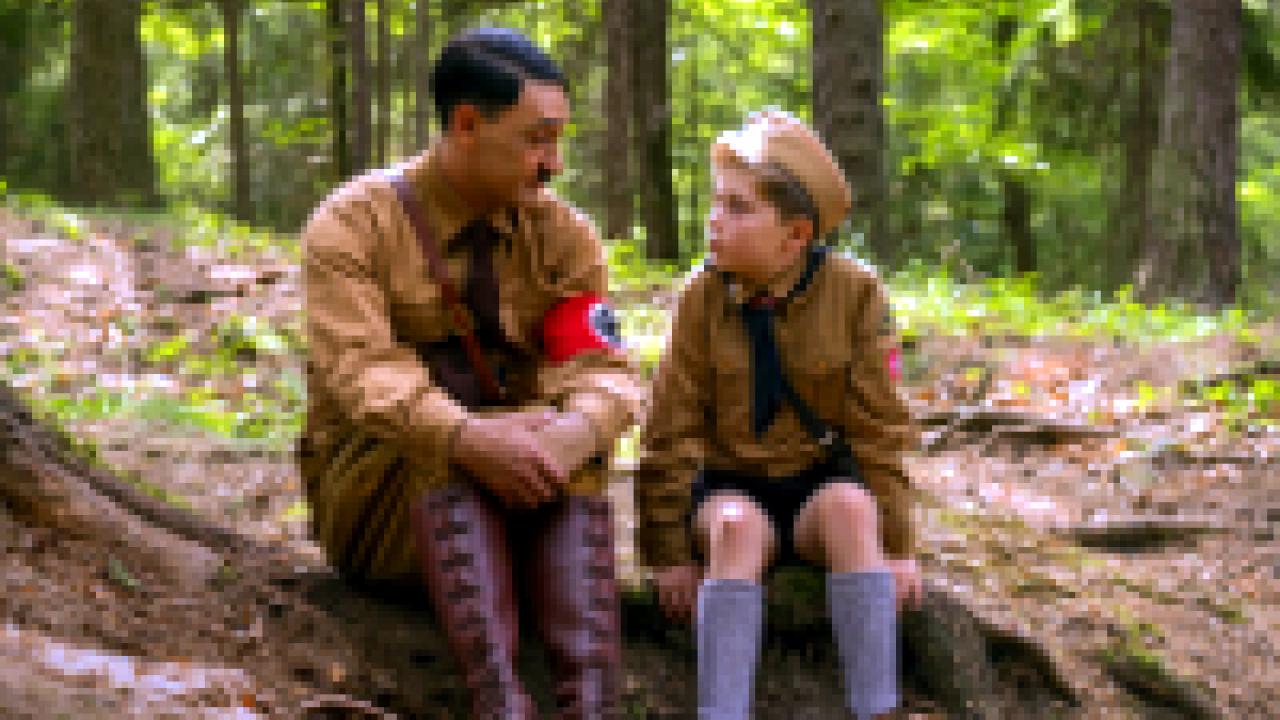 Jojo Rabbit, Szenenbild: Ein Mann, der aussieht wie Adolf Hitler, sitzt im Wald neben einem kleinen Jungen und hört ihm zu.