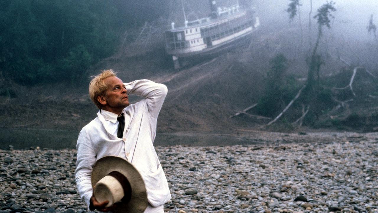 Fitzcarraldo, Szenenbild: Ein Mann im weißen Leinenanzug und mit Hut in der Hand steht in einer Dschungellandschaft und schaut verzweifelt nach oben. Im Hintergrund wird ein Dampfer einen Hang hochgezogen.