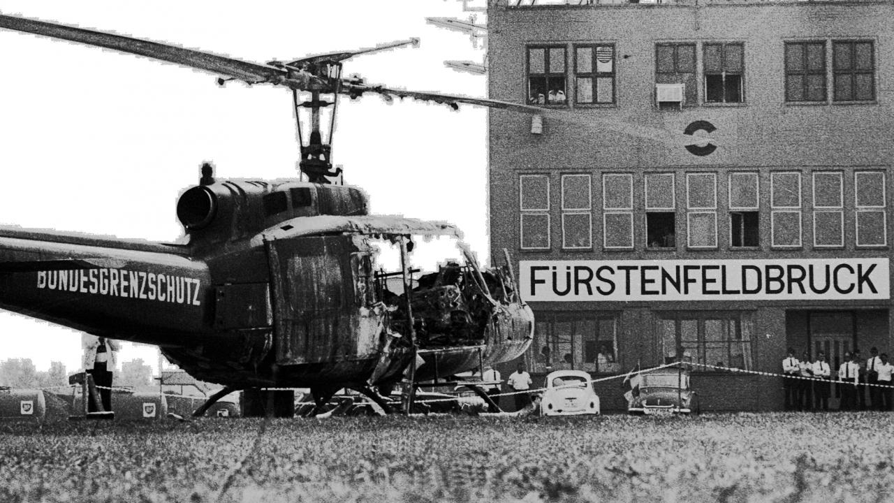 Tod und Spiele - München '72, Szenenbild: Ein ausgebrandter Helicopter mit der Aufschrift "Bundesgrenzschutz" steht vor einem Flughafengebäude mit dem Schriftzug "Fürstenfeldbrück"