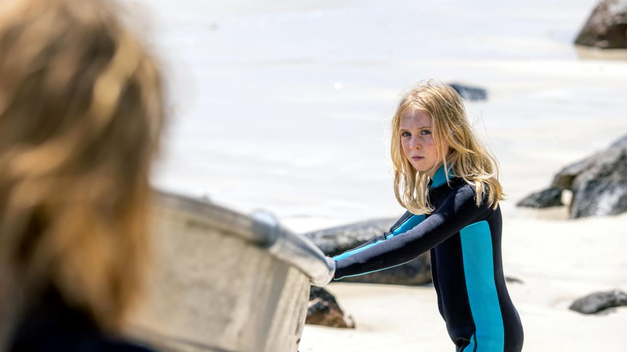 Blueback - Eine tiefe Freundschaft, Szenenbild: Ein Kind steht im Taucheranzug am Strand und hält sich am Rand eines Bootes fest. Es schaut mit ernstem Blick in Richtung einer Person, von der im Vordergrund nur der Hinterkopf unscharf zu erkennen ist