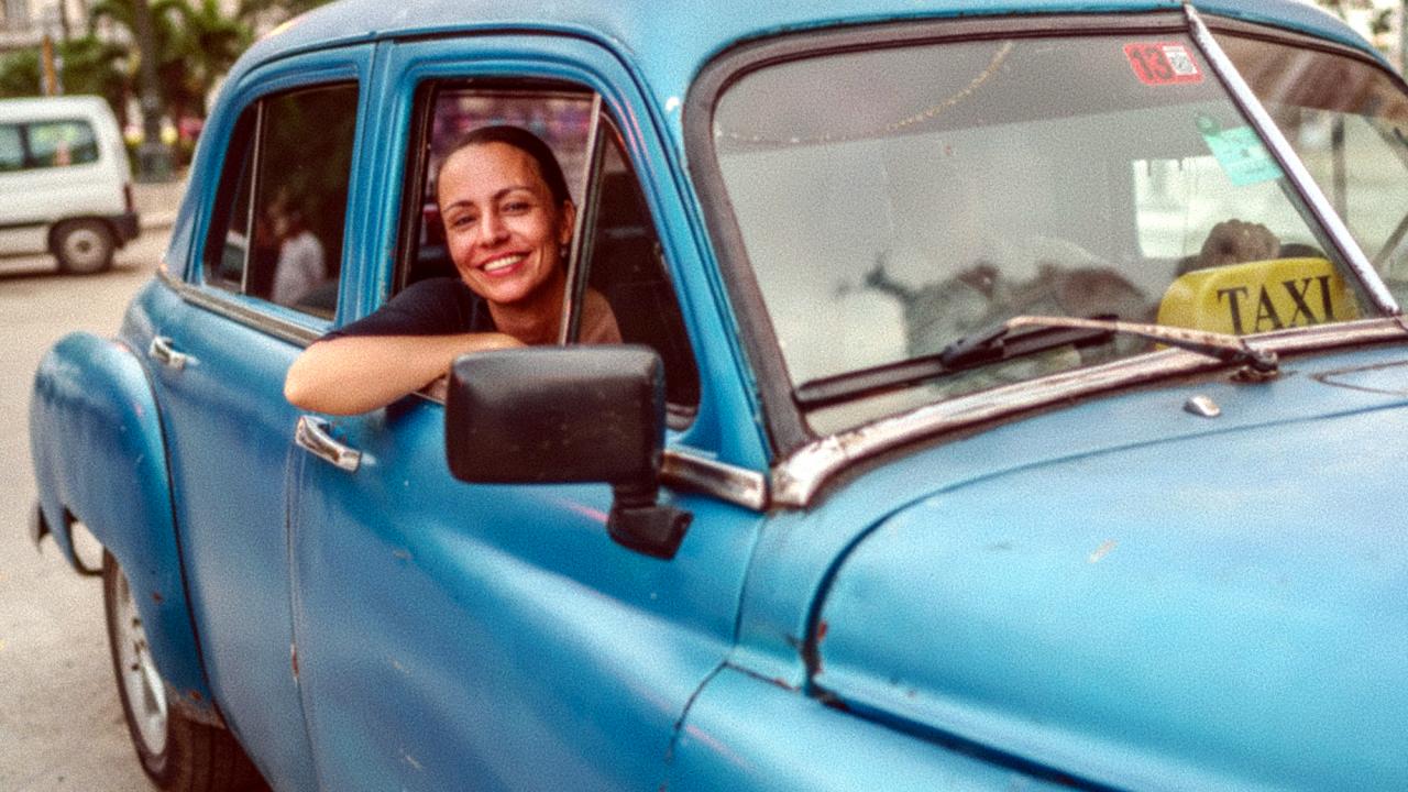 Tanja – Tagebuch einer Guerillera, Szenenbild: Eine junge Frau schaut lächelnd aus dem Seitenfenster eines altmodischen, blauen Taxis.