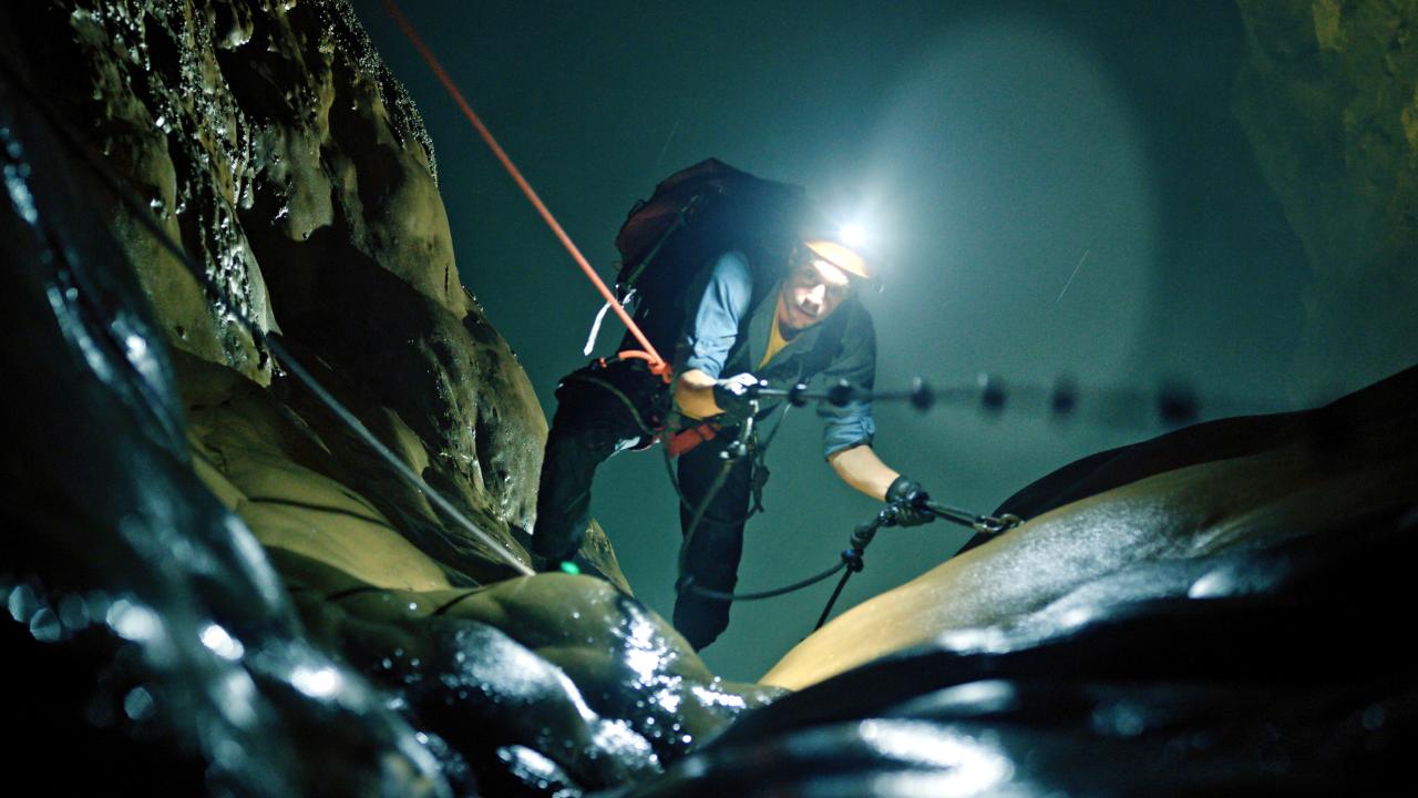 Checker Tobi und die Reise zu den fliegenden Flüssen, Szenenbild: Checker Tobi klettert in einer dunklen Höhle auf die Betrachtenden zu. Er trägt eine Lampe an seinem Helm.