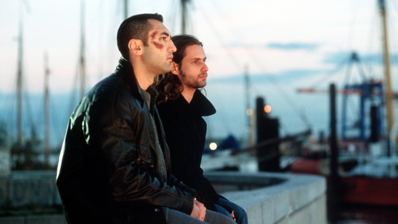 Kurz und schmerzlos, Szenenbild: Zwei junge Männer sitzen nebeneinander auf einer Mauer. Der eine Mann ist im Gesicht verletzt. Im Hintergrund sind Segelboote zu erkennen.