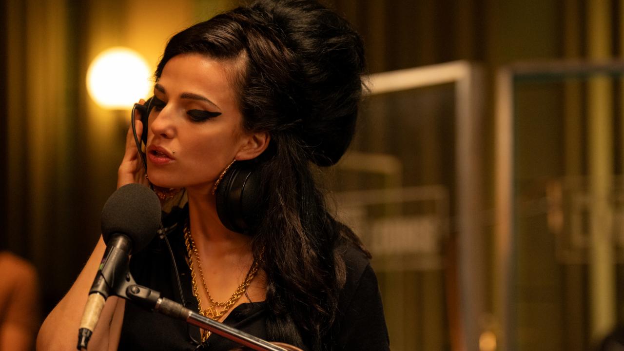 Szenenbild aus dem Film "Back to Black": Die Sängerin Amy Winehouse, dargestellt von Marisa Abela, steht in einem Tonstudio vor einem Mikrofon und singt. Sie hält sich einen Kophörer an ihr rechtes Ohr.