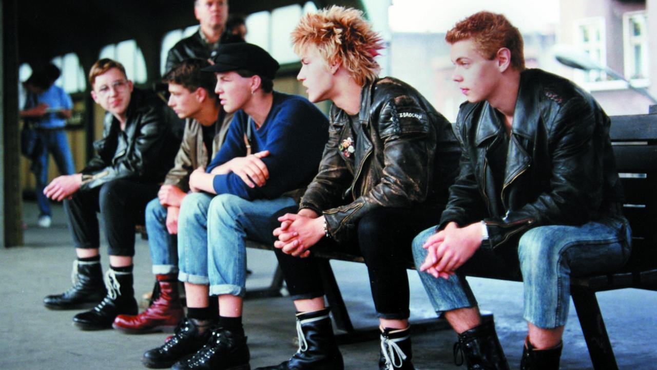 Szenenbild aus dem Dokumentarfilm "flüstern & SCHREIEN": Fünf junge Männer um die 20 sitzen auf einer Bank auf einem U-Bahnhof. Sie tragen Lderjacken, schwere Stiefel oder Punk-Frisuren.