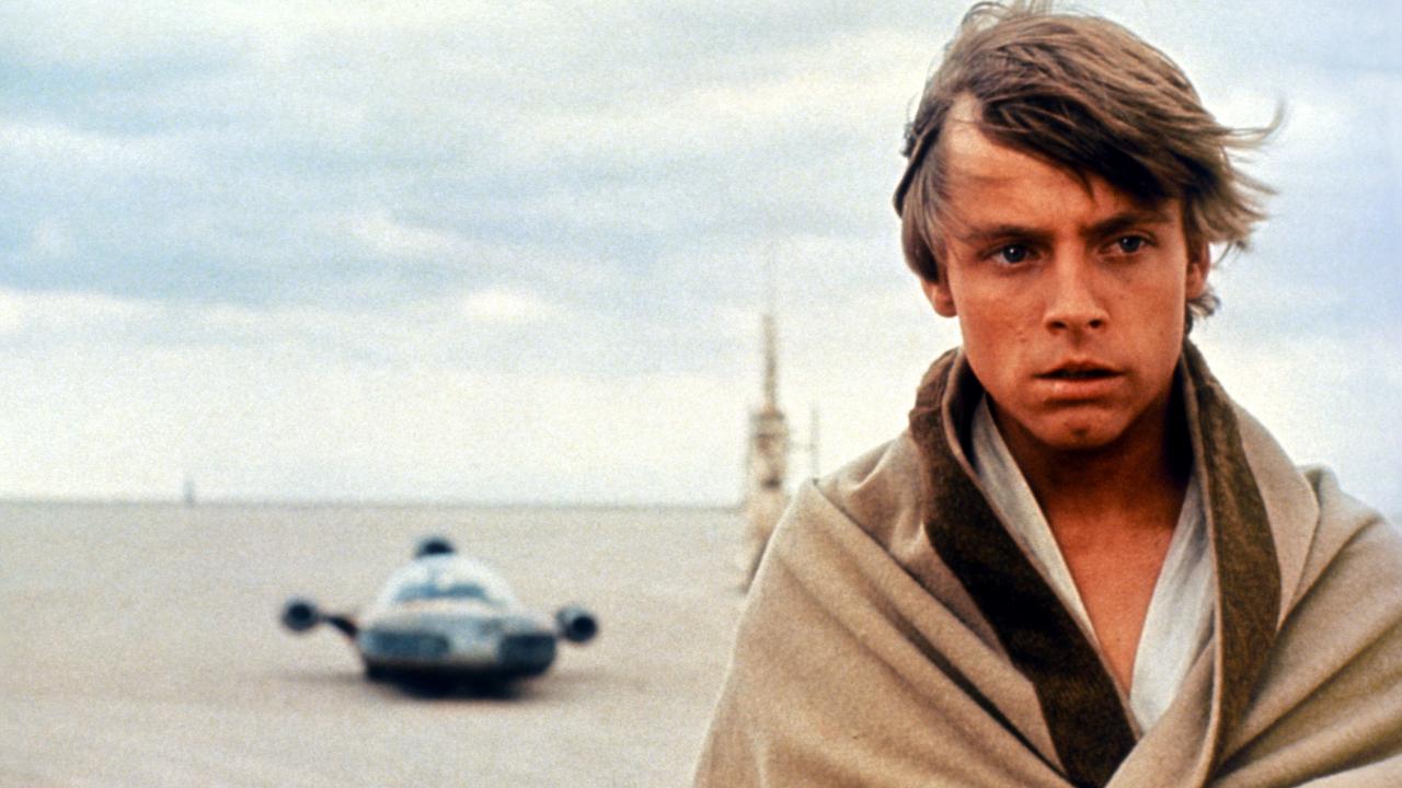 Szenenbild aus dem Film "Star Wars: Episode IV – Eine neue Hoffnung": Nahaufnahme eines jungen Mannes am rechten Bildrand. Er trägt über einem weißen offenen Hemd eine beigefarbene Decke und schaut ernst. Im Hintergrund ist vor fahl-blauem Himmel ein