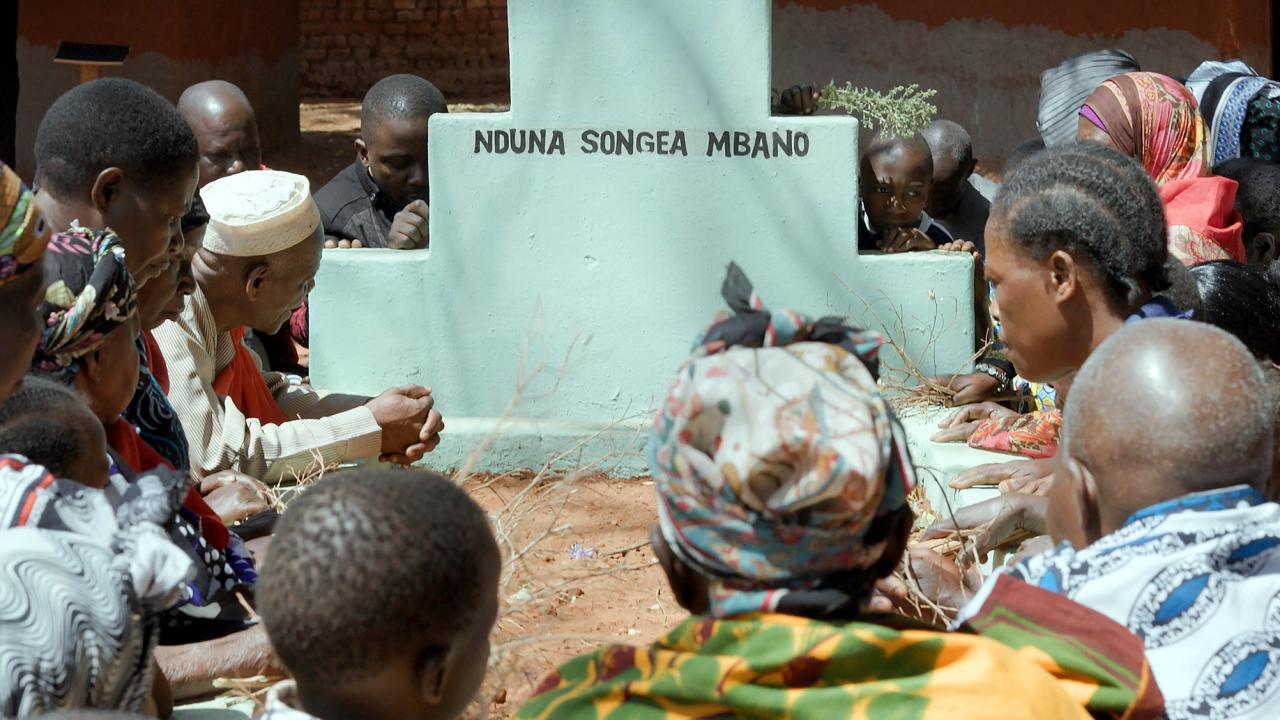 Szenenbild aus dem Dokumentarfilm "Das leere Grab": Nahaufnahme eines Grabstein mit der Inschrift "Nduna Songea Mbano". Um den Stein knien Frauen und Männer. Das Bild wurde in Tansania aufgenommen.