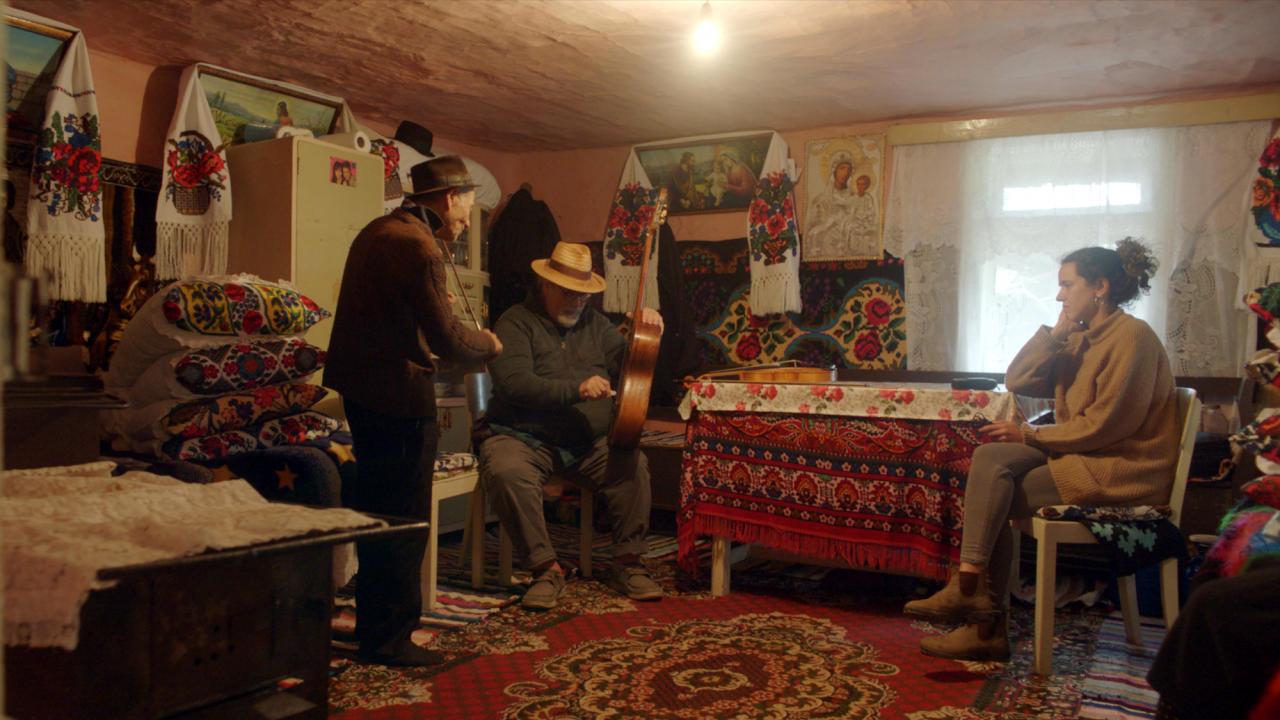 Szenenbild aus dem Film "Das Klezmer Projekt – Eine Reise auf der Suche nach den Wurzeln jiddischer Musik": In einem bunt dekorierten Zimmer musizieren zwei alte Männer auf Gitarre und Violine. Eine jüngere Frau sitzt an einem Tisch und hört zu.