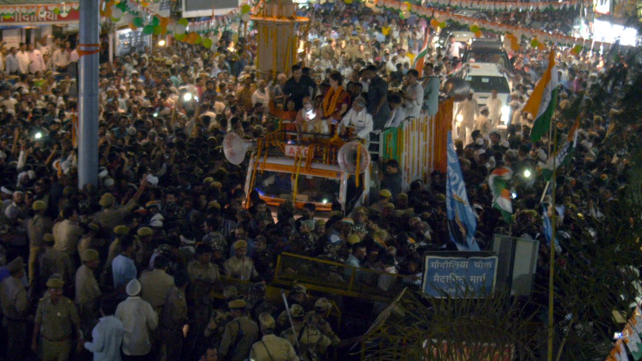 Szenenbild aus dem Dokumentarfilm Don't Worry About India: Dicht gedrängt steht eine Menschenmenge auf einer nächtlichen, mit grün-weiß-orangen Girlanden und Fahnen geschmückten Straße. Sie umgibt ein Autocorso, das von einem