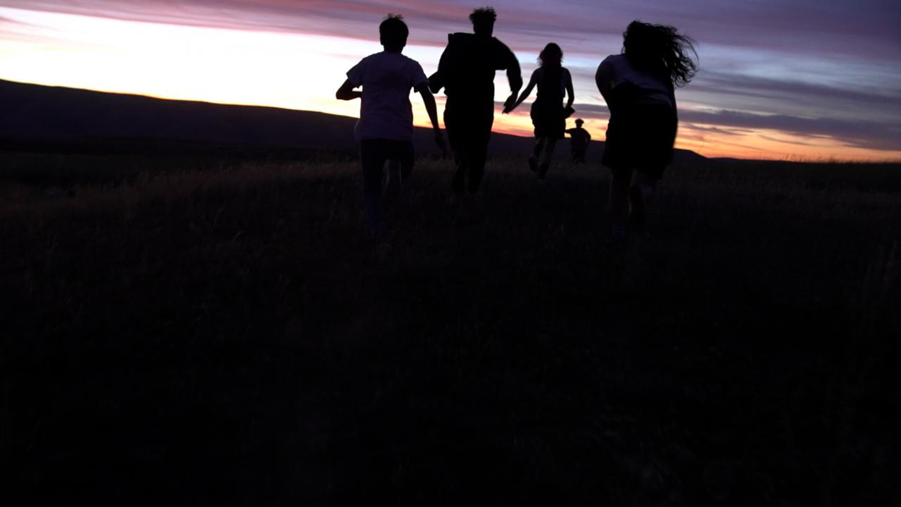 Szenenbild aus dem Roadmovie GASOLINE RAINBOW: Fünf junge Leute laufen in einer Wüstenlandschaft der untergehenden Sonne entgegen. Sie sind nur als schwarze Silhouetten erkennbar.