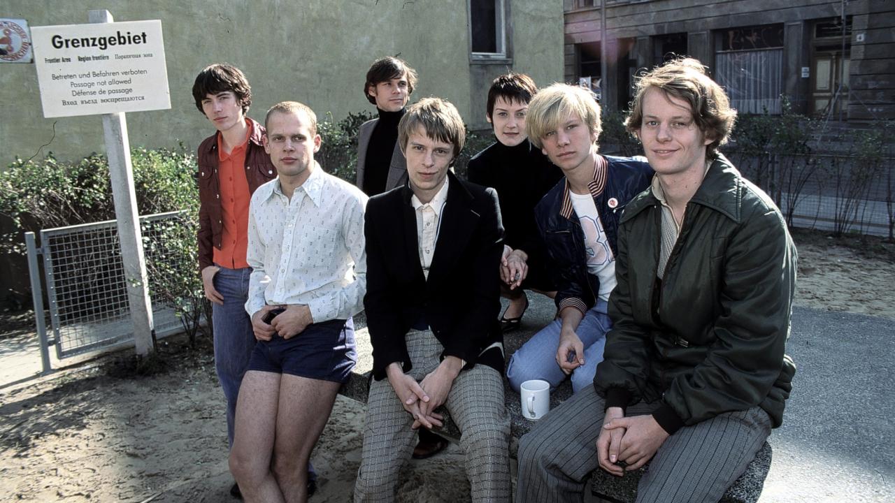 Szenenbild aus dem Film SONNENALLEE: Eine Gruppe von jungen Menschen sitzt auf oder steht neben einem Steintisch auf einem kleinen Platz zwischen mehrgeschossigen Altbauten. Links neben ihnen steht ein Schild mit der Aufschrift "Grenzgebiet. Betreten und