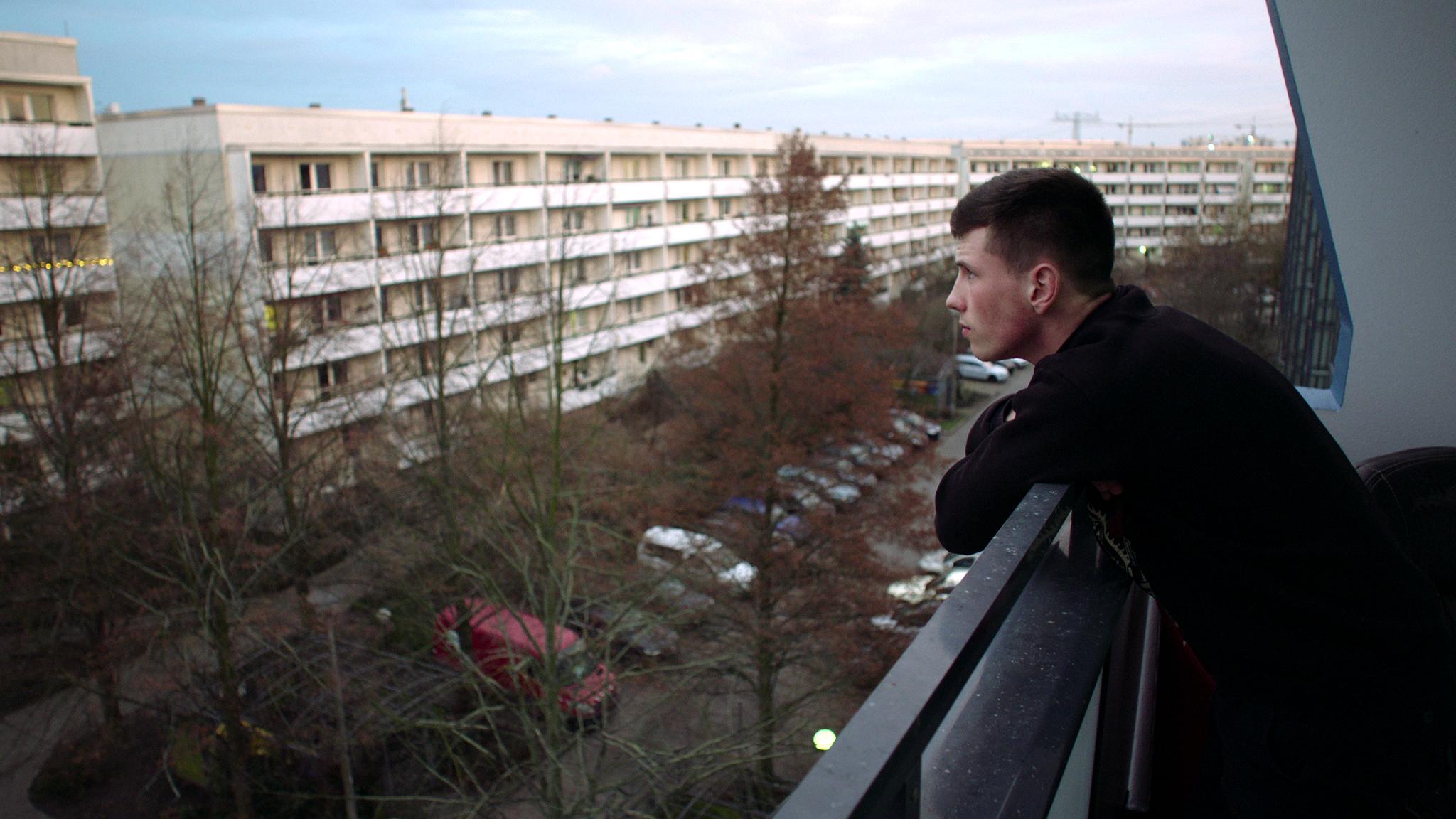 Szenenbild aus dem Dokumentarfilm "Kalle Kosmonaut": Ein junger Mann steht am rechten Bildrand auf einem Balkon. Er schaut auf eine Straße mit Plattenbauten.