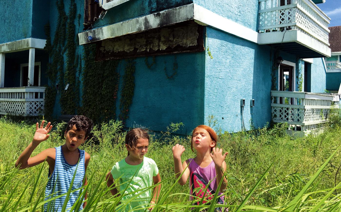 The Florida Project - Szenenbild: Drei Kinder stehen im hohen Gras, dahinter ein türkisfarbenes Haus.