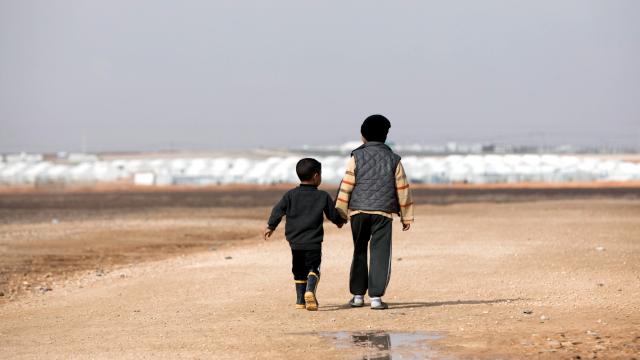 Totale: Zwei Flüchtlingskinder in der Rückenansicht laufen Hand in Hand durch eine karge, sandige Landschaft. Im Hintergrund sind schemenhaft weiße Zelte zu erkennen. Das Foto wurde am 29.01.2018 in Jordanien im Flüchtlingslager Al-Asrak aufgenommen.