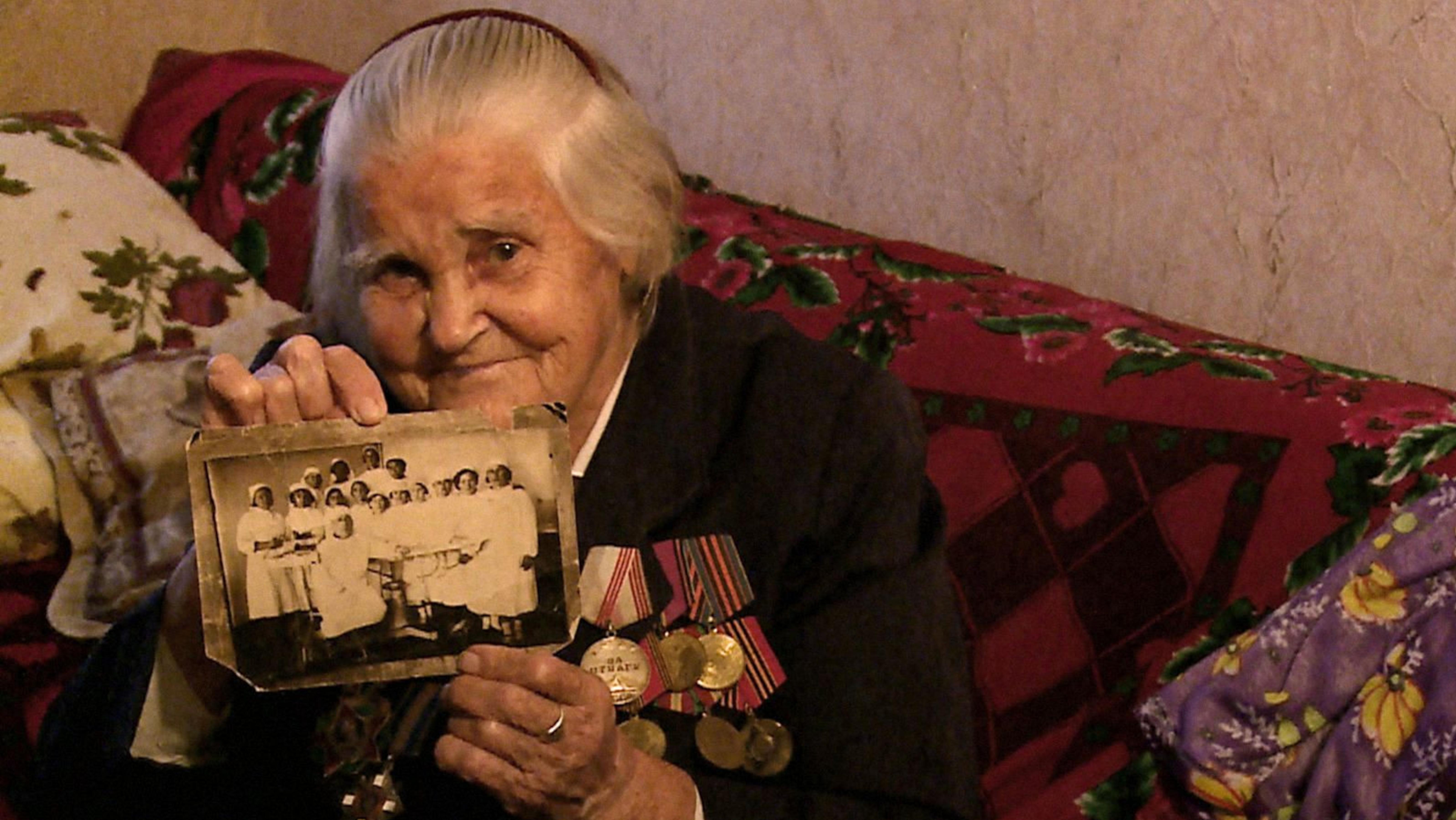 Meine Mutter, ein Krieg und ich, Szenenbild: Eine alte Dame und Veteranin sitzt auf ihrem Sofa und hält lächelnd eine Schwarz-Weiß-Fotografie in die Kamera