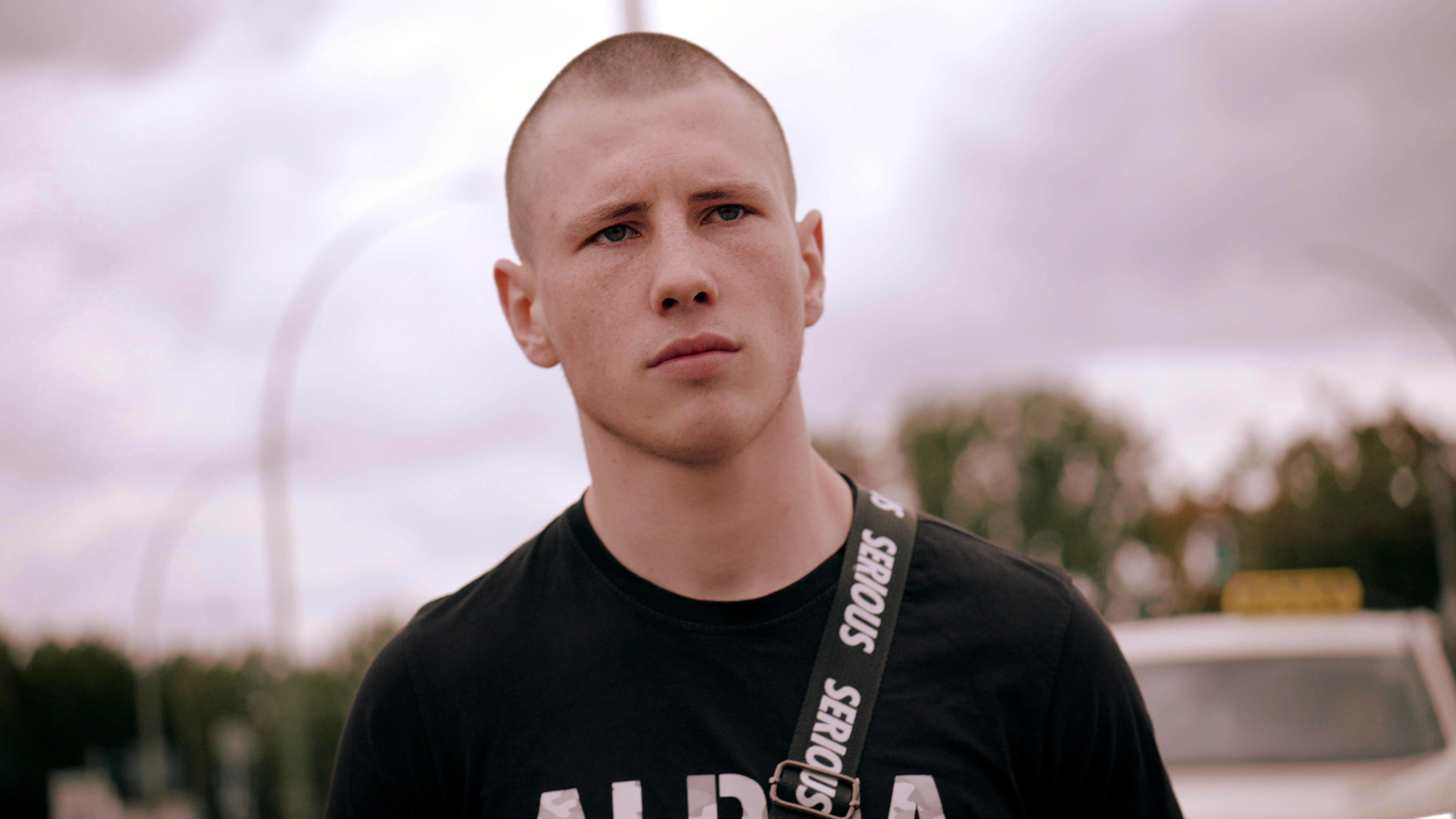 Kalle Kosmonaut, Szenenbild: Nahe Porträtaufnahme eines jungen Mannes mit kahl geschorenem Kopf und in schwarzem T-Shirt. Im Hintergrund ist ein wolkiher Himmel zu sehen.