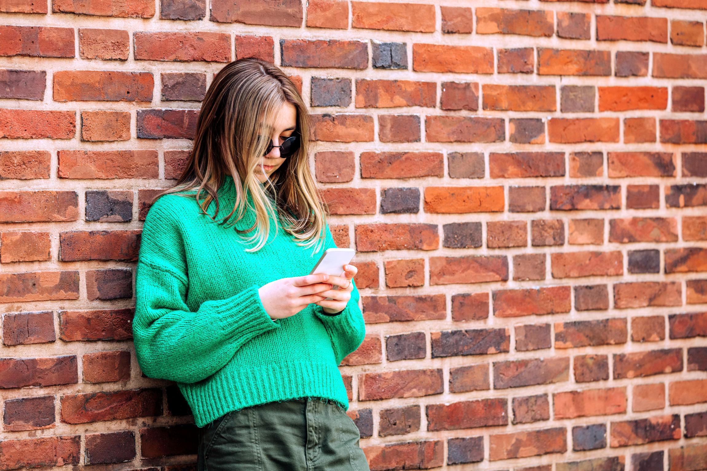 Ein Mädchen im Teenageralter lehnt an einer Mauer und schaut auf ihr Handy