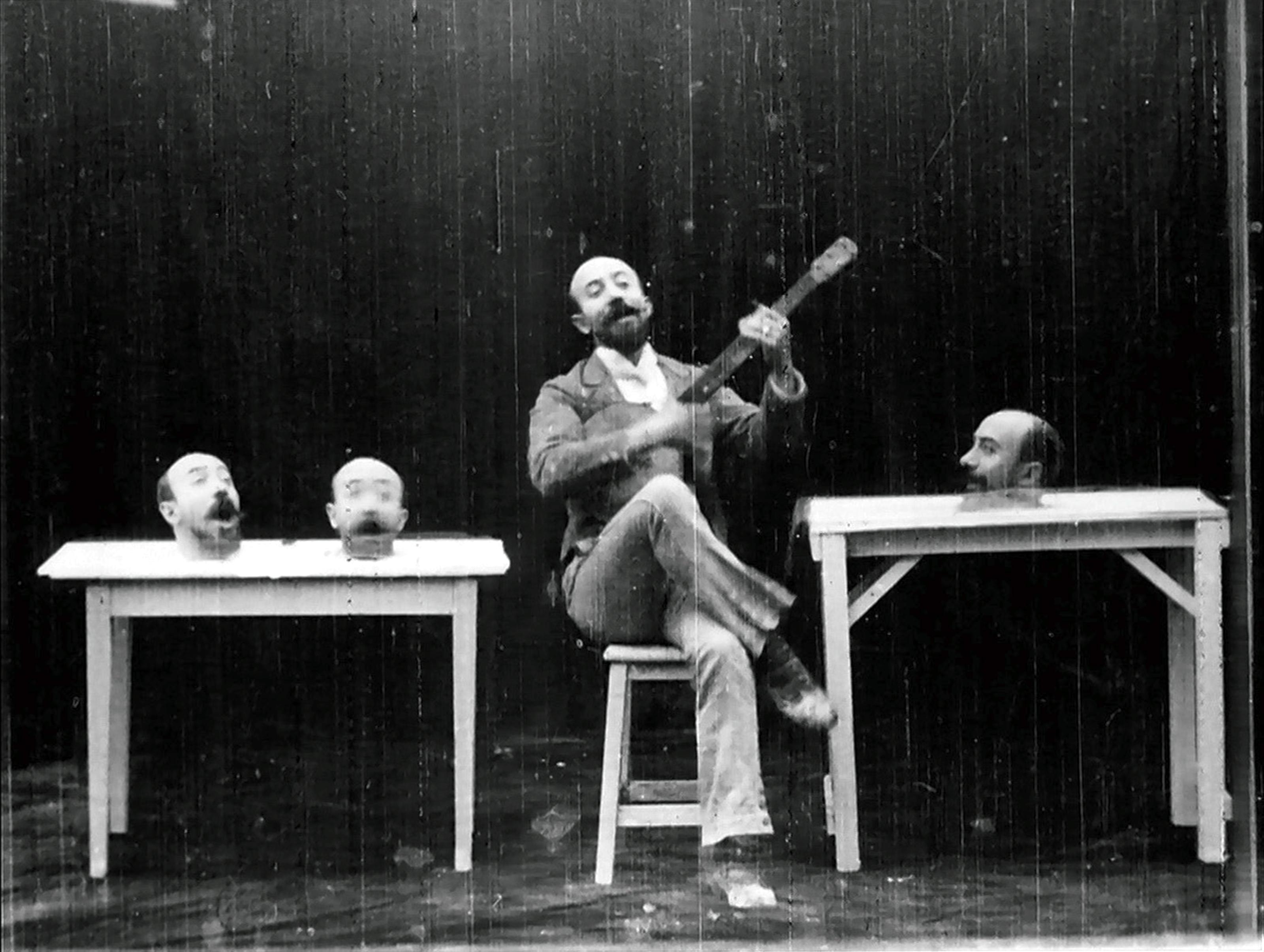 Szenenbild aus dem Film Un homme de têtes von Georges Méliès (1898): Ein Mann sitzt mit einer Gitarre auf einem Stuhl. Auf Tischen neben ihm liegen drei Köpfe von ihm, die singen.