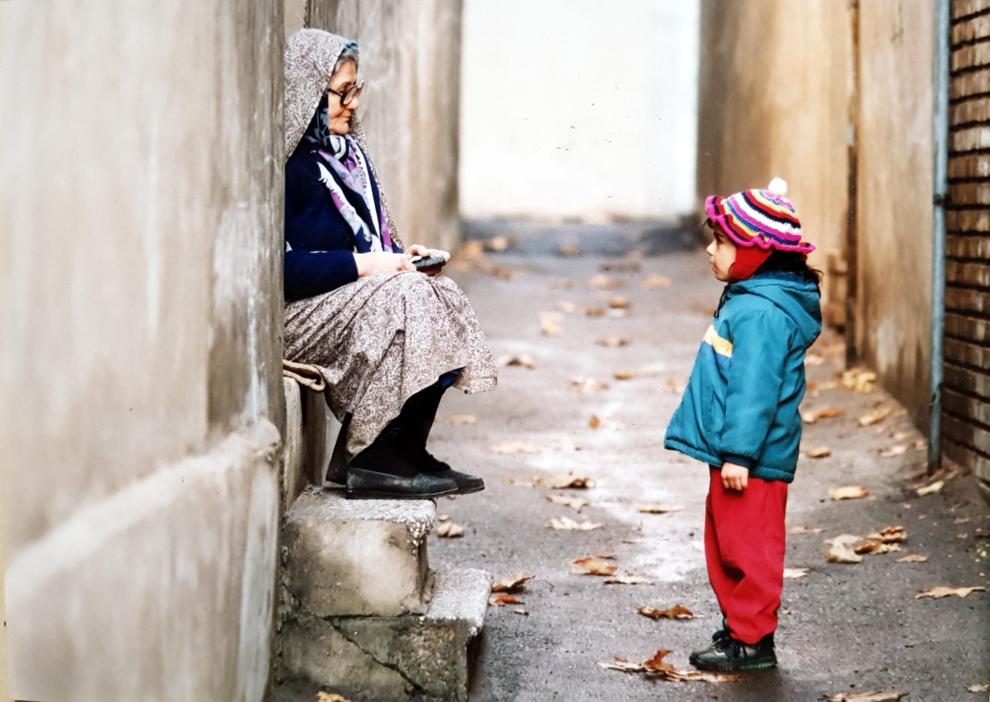 Ein Sack Reis, Szenenbild: In einer engen Gasse sitzt eine alte Frau am linken Bildrand auf der Stufe einer Tür. Ihr gegenüber steht ein Kind. Beide schauen sich an. (Foto: Mitra Mahasani, Produzent: Mohammad Ali Talebi.)