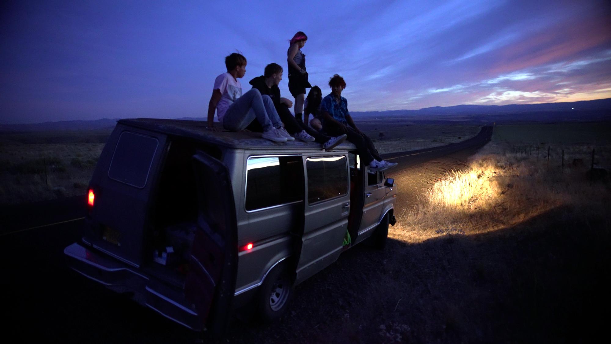 Szenenbild aus dem Roadmovie GASOLINE RAINBOW: Drei junge Männer und zwei junge Frauen stehen auf dem Dach eines Campervans. Die Sonne geht unter.